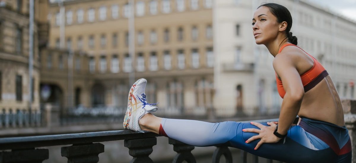 Woman stretching outdoors on urban street railing in activewear, promoting fitness.