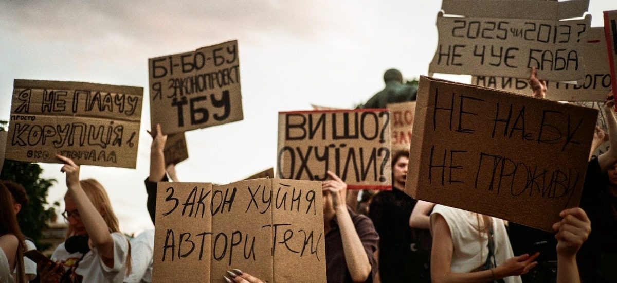 Protesters hold signs with handwritten messages against a cloudy sky.