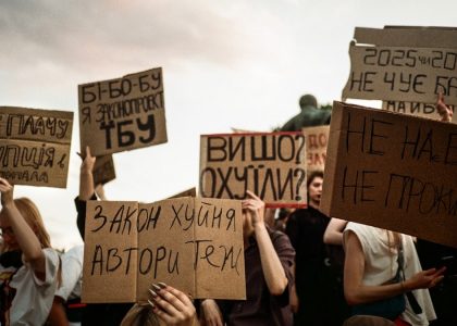Protesters hold signs with handwritten messages against a cloudy sky.